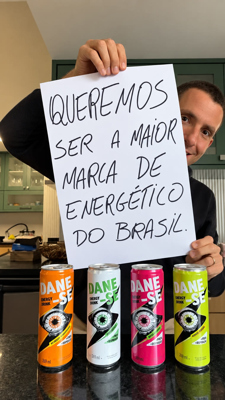 O fundador da Dane-se segurando um cartaz escrito à mão: queremos ser a maior marca de energético do Brasil, com as quatro latas na bancada da cozinha.